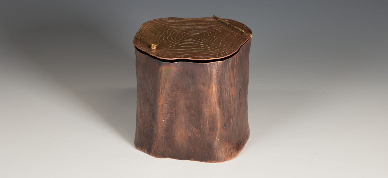 A copper and bronze stump sculpture. It has a handle and hinge on top.