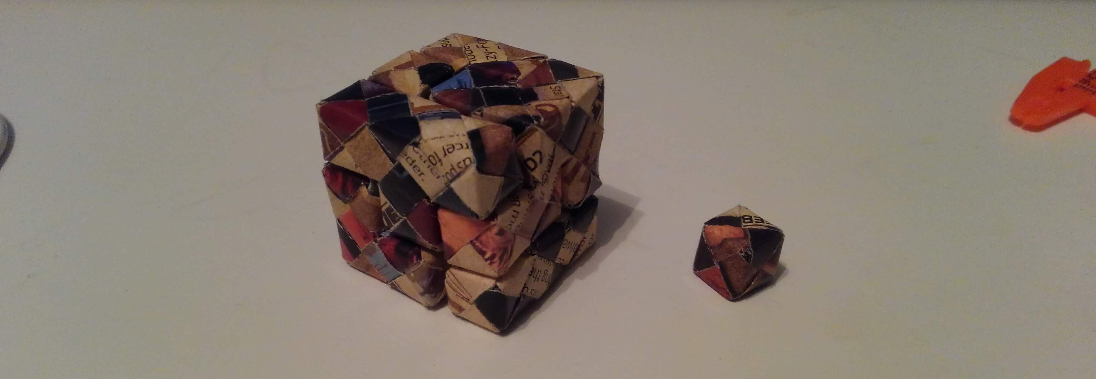 An origami sonobe puzzle cube standing next to a standard sonobe cube. It is 3x longer in each dimension.